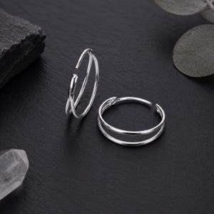 Puede incluir: Dos anillos de plata para el dedo gordo del pie sobre una superficie negra. Uno tiene un diseño de doble banda, mientras que el otro presenta un diseño único y superpuesto. La joyería es simple y elegante, adecuada para el uso diario.