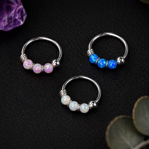 Può includere: Tre anelli circolari in metallo, ciascuno caratterizzato da tre piccole perline sferiche e una perlina in metallo liscia. Un anello ha perline rosa, uno ha perline blu e uno ha perline bianche. Gli anelli sono disposti in una forma triangolare.