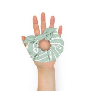 May include: A sage green scrunchie featuring a white abstract line pattern and a decorative bow tie detail. The fabric is gathered around an elastic band, creating a ruffled texture. The design consists of continuous white lines on a solid green background.