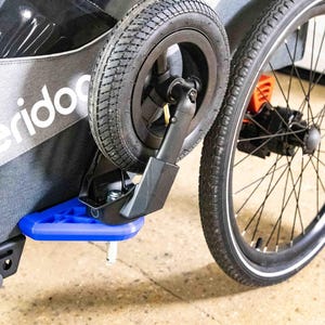Op de afbeelding: Close-up van een zwart en grijs fietsaanhangerwiel met een zwarte band. De aanhanger heeft een blauwe voetensteun en de tekst "SPORTREX" is zichtbaar. De aanhanger is ontworpen voor buitenactiviteiten.