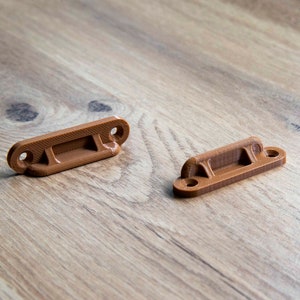 Może przedstawiać: Trzy brązowe, prostokątne wsporniki z zaokrąglonymi końcami i dwoma otworami każdy są ułożone na drewnianej powierzchni. Wsporniki mają podniesioną środkową część, prawdopodobnie do montażu lub podparcia.