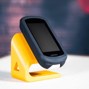 Könnte beinhalten: Ein schwarzes Garmin-Gerät in einem dunkelblauen Gehäuse wird auf einem leuchtend gelben Ständer präsentiert. Der Ständer hat einen dreieckigen Ausschnitt. Das Garmin-Gerät hat einen schwarzen Bildschirm und das Garmin-Logo ist sichtbar. Der Hintergrund ist unscharf.