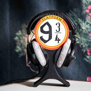 Peut inclure: Casque audio noir avec coussinets gris posé sur un support noir. Un panneau circulaire avec les mots "PLATFORM" et les chiffres "9 3/4" est centré au-dessus du casque. L'arrière-plan est sombre avec un soupçon de verdure.