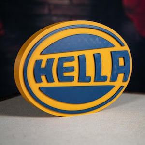 Peut inclure: Un panneau rond jaune et bleu avec le mot "HELLA" en lettres bleues. Le panneau a un aspect en relief et tridimensionnel, sur un fond flou.