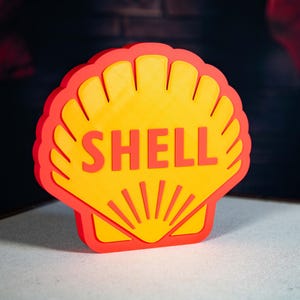 Op de afbeelding: Een driedimensionaal Shell-logo in de vorm van een schelp. Het logo heeft een gele binnenkant met het woord 'SHELL' in oranje en een rode rand. De afbeelding is goed belicht en toont de kleuren en het ontwerp.