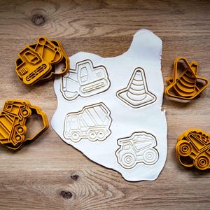 Könnte beinhalten: Gelbe Ausstecher aus Kunststoff in Form von Baumaschinen und einem Leitkegel. Die Ausstecher sind um ein Stück weißen Teigs mit passenden Fahrzeugumrissen angeordnet. Der Hintergrund ist eine Holzoberfläche.