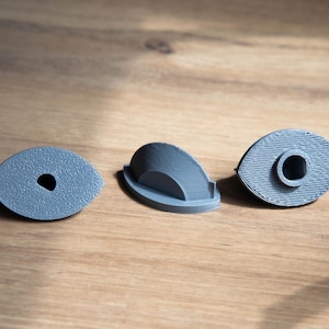Peut inclure: Trois objets gris imprimés en 3D sont disposés sur une surface en bois. Les objets comprennent deux pièces ovales avec des trous et une pièce incurvée semi-circulaire. Les objets semblent être des composants d'un élément plus grand.