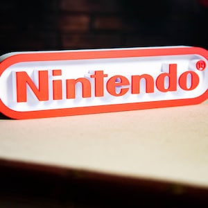 Peut inclure: Logo Nintendo blanc et rouge, avec le mot "Nintendo" en rouge sur fond blanc. Le logo a une forme rectangulaire arrondie avec un bord rouge. Le fond est de couleur beige clair.