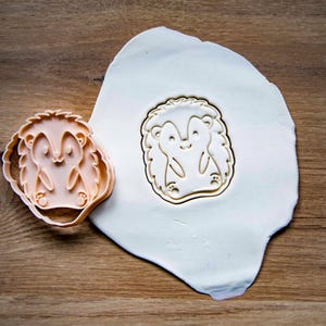 May include: A light orange hedgehog-shaped cookie cutter next to a pressed impression of the same design in white dough. The cutter is on a wooden surface, and the dough is on a white surface.