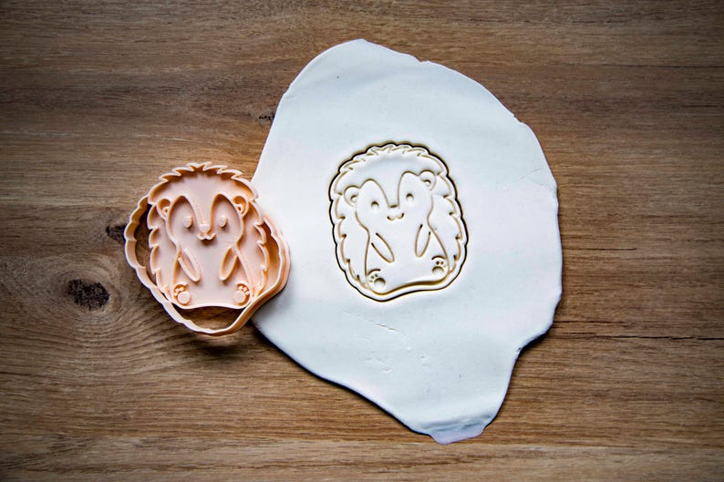 May include: A light orange hedgehog-shaped cookie cutter next to a pressed impression of the same design in white dough. The cutter is on a wooden surface, and the dough is on a white surface.