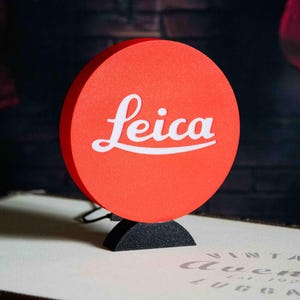 Peut inclure: Un panneau rond rouge avec le mot "Leica" en blanc. Le panneau est monté sur une base noire semi-circulaire. L'arrière-plan est sombre, avec un soupçon de chaussette de Noël.