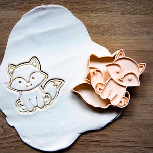 May include: Two fox-shaped cookie cutters, one pressed into white dough, the other unpressed. The cutters are light orange and feature a smiling fox design. The dough sits on a wooden surface, suggesting a baking theme.