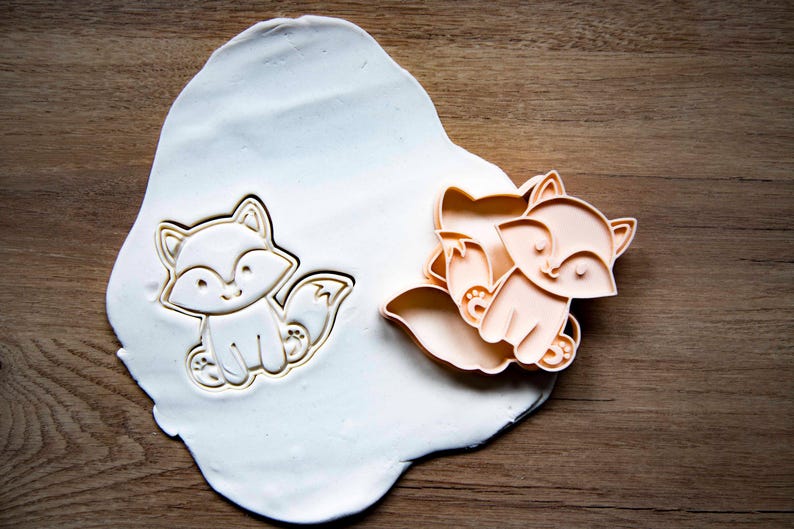 May include: Two fox-shaped cookie cutters, one pressed into white dough, the other unpressed. The cutters are light orange and feature a smiling fox design. The dough sits on a wooden surface, suggesting a baking theme.