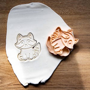 May include: A raccoon-shaped cookie cutter and a fox-shaped cookie cutter are displayed on a white surface. The cutters are made of a light orange plastic and are sitting on a white surface. The background is a wooden surface.