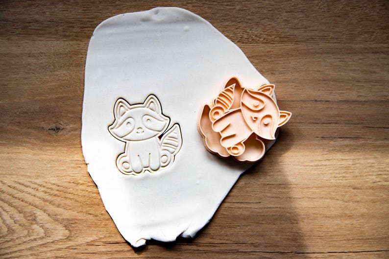 May include: A raccoon-shaped cookie cutter and a fox-shaped cookie cutter are displayed on a white surface. The cutters are made of a light orange plastic and are sitting on a white surface. The background is a wooden surface.