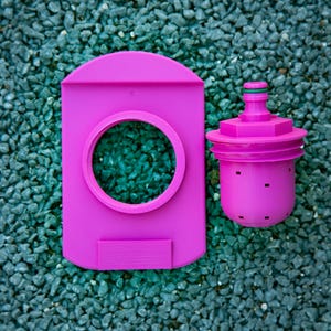 May include: A bright pink water filter system, featuring a rectangular housing with a circular opening and a separate cylindrical filter component. The filter has a green accent and small square perforations. The items are set against a background of small, dark gray stones.