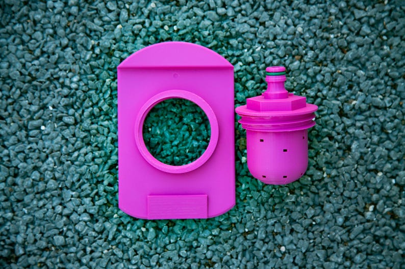 May include: A bright pink water filter system, featuring a rectangular housing with a circular opening and a separate cylindrical filter component. The filter has a green accent and small square perforations. The items are set against a background of small, dark gray stones.