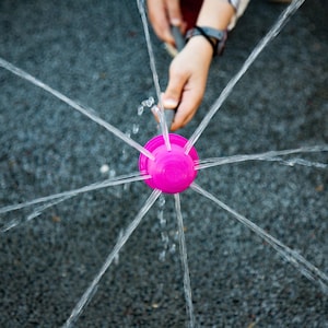 May include: A bright pink water sprinkler sprays water in multiple directions. The sprinkler is held by a person's hand, creating a playful outdoor water feature. The water streams create a radial pattern.
