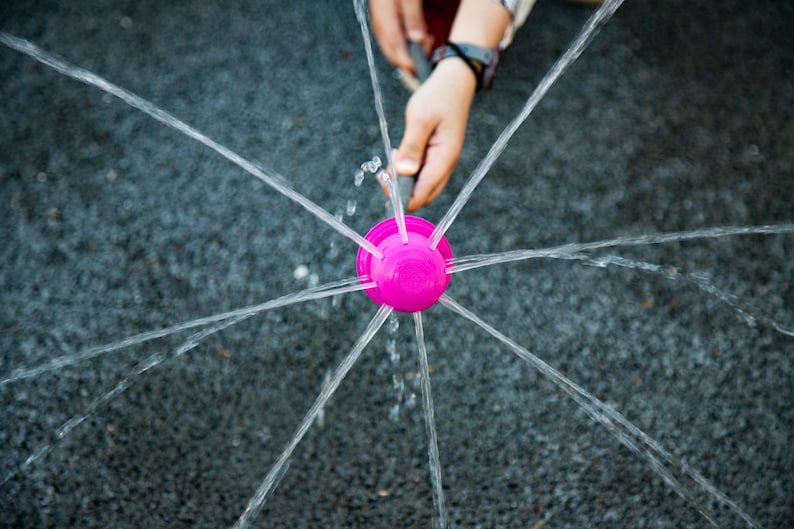 May include: A bright pink water sprinkler sprays water in multiple directions. The sprinkler is held by a person's hand, creating a playful outdoor water feature. The water streams create a radial pattern.