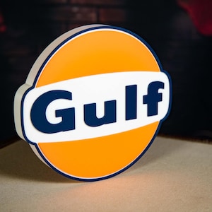 Pode incluir: Uma placa redonda com a palavra "Gulf" em letras azul escuro. A placa tem um centro laranja, uma faixa branca e uma borda azul escuro. É um logotipo de estilo vintage.