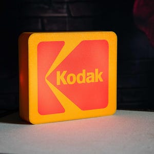 Könnte beinhalten: Ein rechteckiges, gelb-rotes Produkt mit dem Kodak-Logo. Das Kodak-Logo ist in gelbem Text auf rotem Hintergrund dargestellt. Das Produkt hat abgerundete Ecken und ist vor einem dunklen Hintergrund platziert.