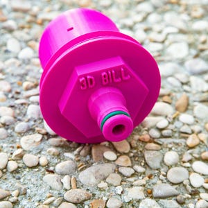 May include: A bright pink 3D-printed object with the text "3D BILL" on its hexagonal base. The object has a cylindrical body and a nozzle with a green ring. It rests on a textured, stone surface.