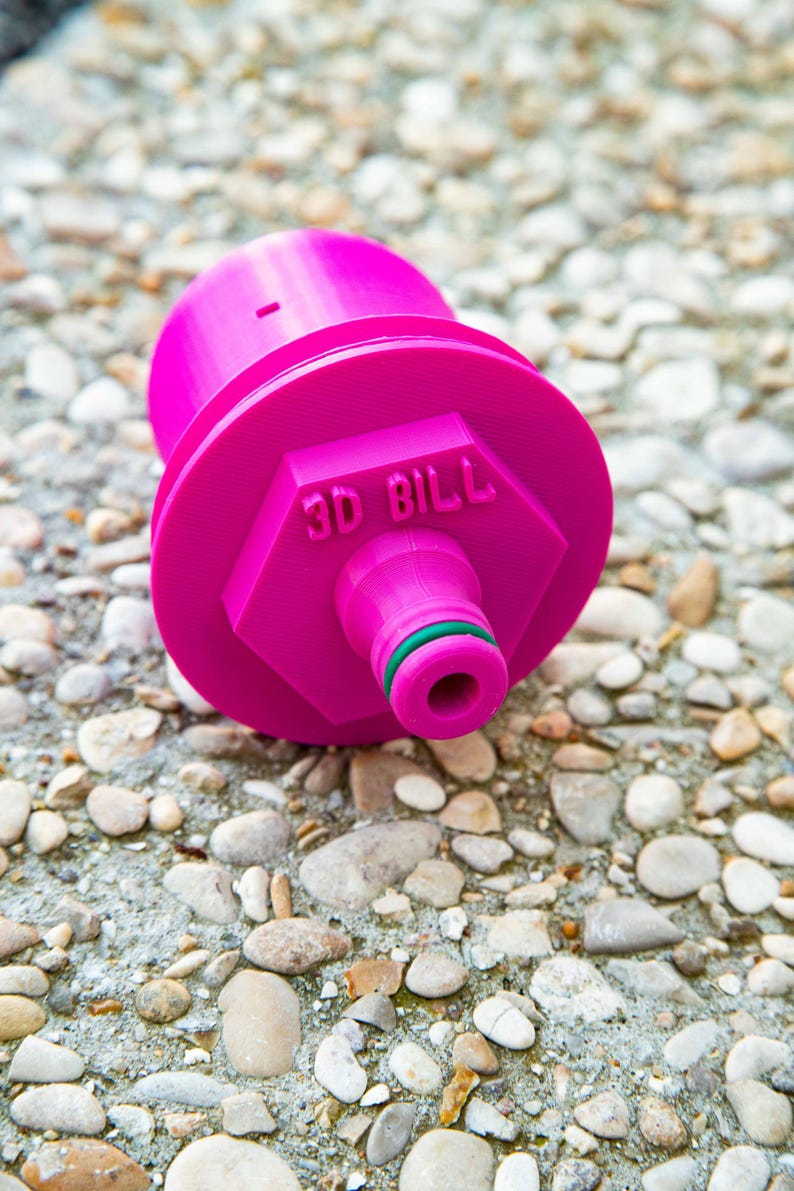 May include: A bright pink 3D-printed object with the text "3D BILL" on its hexagonal base. The object has a cylindrical body and a nozzle with a green ring. It rests on a textured, stone surface.