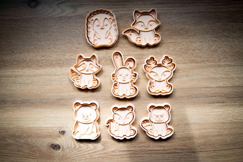 May include: A collection of eight light brown animal-shaped cookie cutters. The set includes a hedgehog, fox, raccoon, rabbit, deer, bear, squirrel, and beaver. The cutters are arranged on a wooden surface.