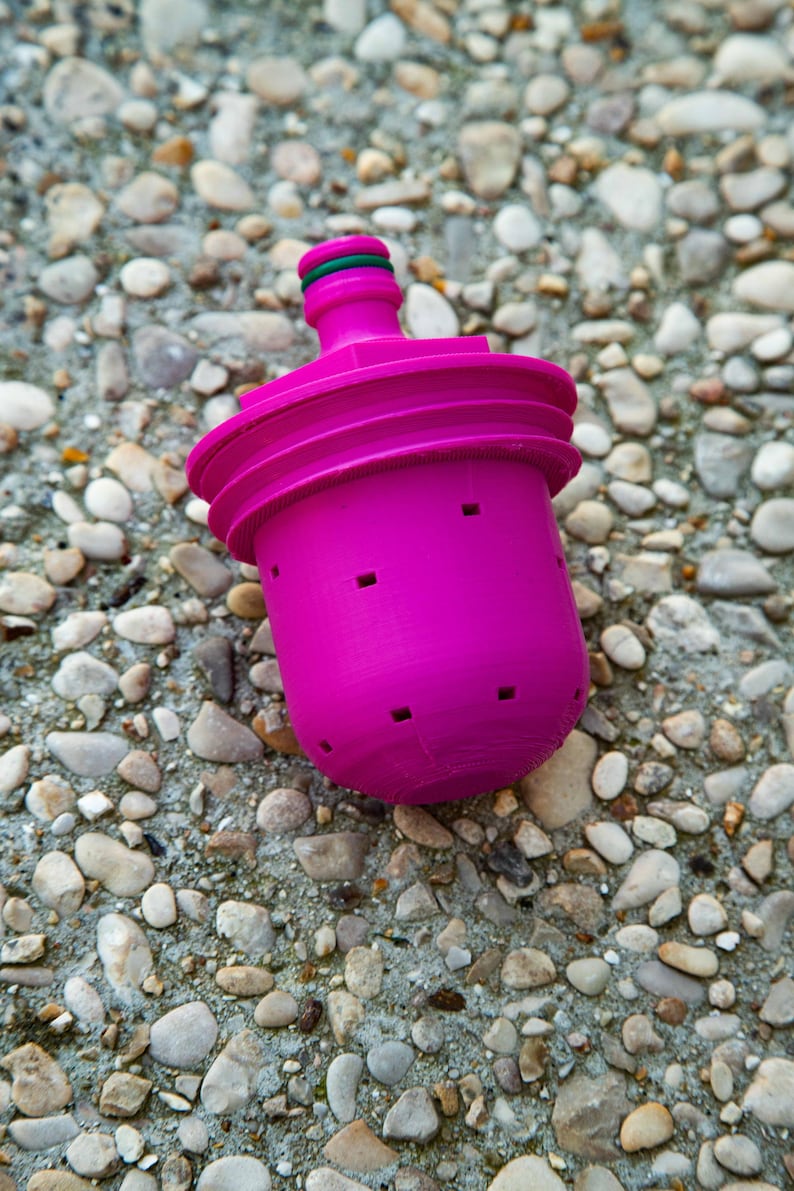 May include: A bright magenta water filter with a green O-ring. The filter has a cylindrical body with several small square holes and a ridged top. The filter is sitting on a surface of small pebbles.