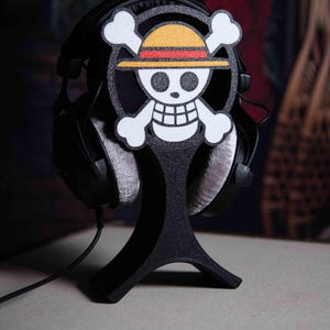 Op de afbeelding: Zwarte hoofdtelefoonstandaard met het One Piece Jolly Roger-logo. De standaard heeft een zwarte basis en een cirkelvormig frame met het iconische schedel- en kruisbeendesign. De hoofdtelefoon is zwart met grijze oorschelpen.