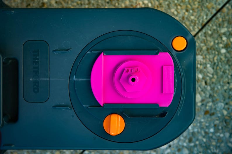 May include: A close-up of a dark gray plastic component with a bright pink central piece. The component has two orange circular buttons and the text "THETFORD" is visible. The design is geometric and functional.