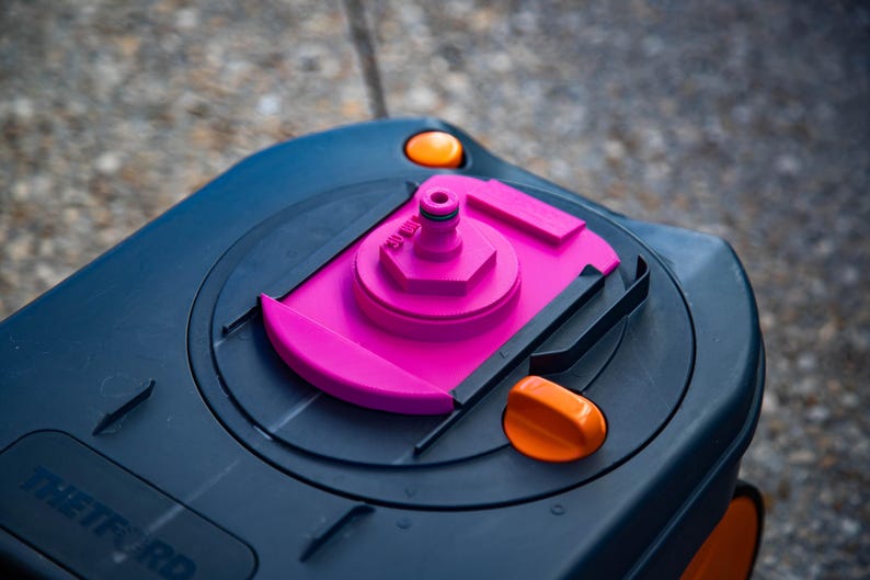 May include: Close-up of a dark blue RV water tank lid with a bright pink fill port and orange accents. The lid has a rounded shape with a textured surface and is designed for outdoor use.
