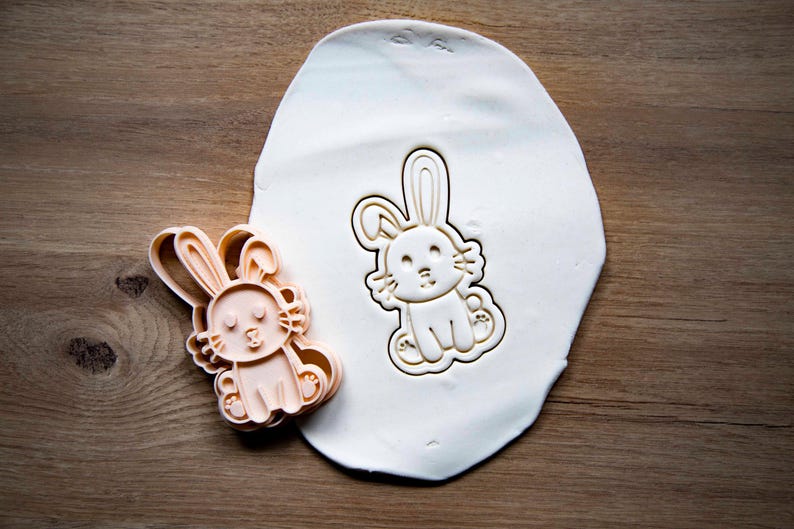 May include: A light peach-colored bunny-shaped cookie cutter next to a bunny-shaped imprint in white dough. The cutter has a detailed design of a bunny with long ears, a smiling face, and seated pose. The dough is on a wooden surface.