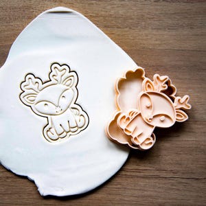 May include: A light-colored, seated deer cookie cutter and its impression in a white dough. The cutter is a light orange color. The dough is on a wooden surface. The deer has large antlers and a friendly face. The cookie cutter is for baking.