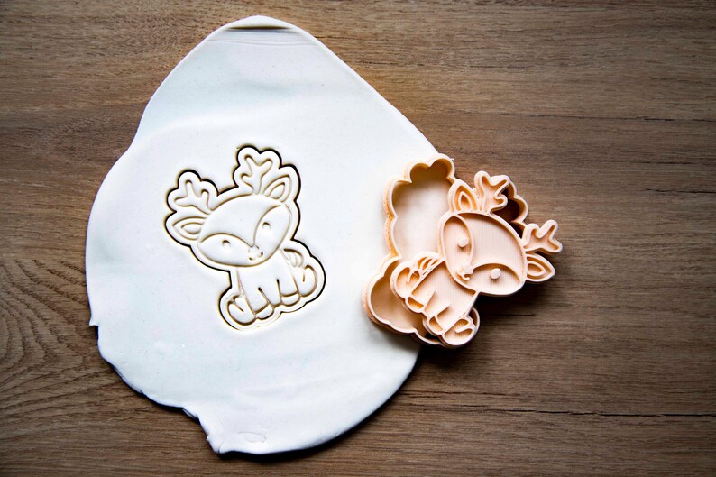 May include: A light-colored, seated deer cookie cutter and its impression in a white dough. The cutter is a light orange color. The dough is on a wooden surface. The deer has large antlers and a friendly face. The cookie cutter is for baking.