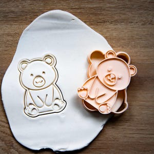 May include: A bear-shaped cookie cutter and a bear-shaped cookie are shown on a wooden surface. The cookie cutter is peach-colored, and the cookie is white. The bear is sitting with its legs forward.