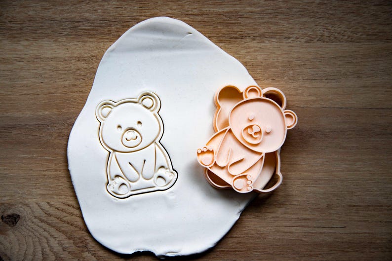 May include: A bear-shaped cookie cutter and a bear-shaped cookie are shown on a wooden surface. The cookie cutter is peach-colored, and the cookie is white. The bear is sitting with its legs forward.