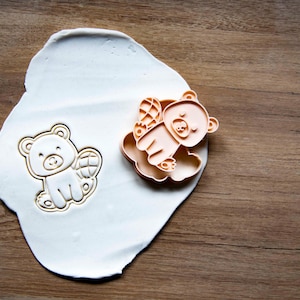 May include: A bear-shaped cookie cutter and a bear-shaped cookie on a white surface, next to a wooden surface. The cookie cutter is light orange, and the cookie is light beige. The bear has wings.