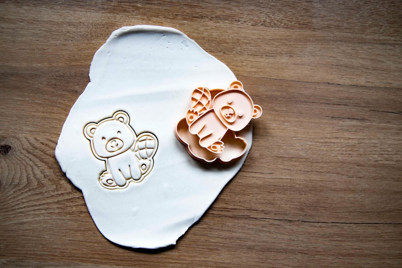 May include: A bear-shaped cookie cutter and a bear-shaped cookie on a white surface, next to a wooden surface. The cookie cutter is light orange, and the cookie is light beige. The bear has wings.
