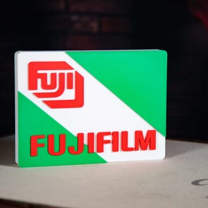 May include: A rectangular Fujifilm sign featuring a white diagonal stripe, green background, and red logo and text. The sign is a promotional item for the Fujifilm brand.