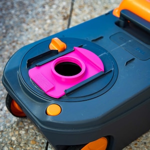 May include: A dark gray portable toilet with orange accents, including wheels, a handle, and buttons. The toilet features a bright pink opening and is designed for outdoor use. The image is taken from a high angle.