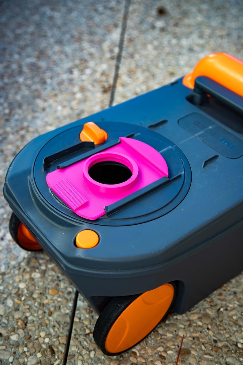 May include: A dark gray portable toilet with orange accents, including wheels, a handle, and buttons. The toilet features a bright pink opening and is designed for outdoor use. The image is taken from a high angle.
