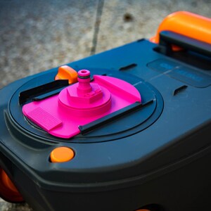 May include: A close-up of a black and gray portable toilet cassette with orange and pink accents. The cassette has a pink water inlet and an orange handle. The wheels are orange.