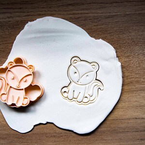May include: Two fox-shaped cookie cutters. One is a peach color, the other is a white outline. Both are on a white surface, set against a wooden background. The cutters are designed with a cute, cartoonish style.