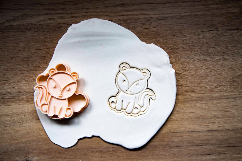 May include: Two fox-shaped cookie cutters. One is a peach color, the other is a white outline. Both are on a white surface, set against a wooden background. The cutters are designed with a cute, cartoonish style.
