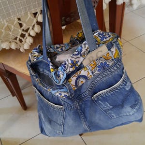May include: A blue denim tote bag constructed from repurposed jeans, featuring two shoulder straps and exterior pockets. The top opening is lined with a fabric band displaying a blue, yellow, and white floral pattern, with the letters RC visible.