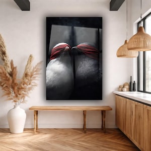 May include: A framed art print featuring two birds with red blindfolds, facing each other. One bird is white, the other is gray. The artwork is displayed above a wooden bench in a room with a vase of dried plants.