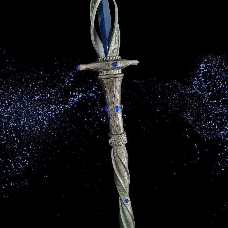 May include: A silver wand with a blue crystal at the top and blue gems along the shaft. The wand is against a black background with blue glitter.