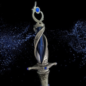 May include: A silver-toned decorative sword with a blue crystal and blue gem accents. The sword has a detailed design with intricate swirls and a pointed tip.