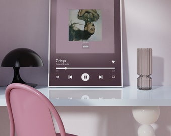 May include: A framed print of a music album cover featuring a woman with long dark hair lying on her back. The album cover is for the song "7 rings" by Ariana Grande. The print is on a white desk with a lamp, a vase, and a chair.